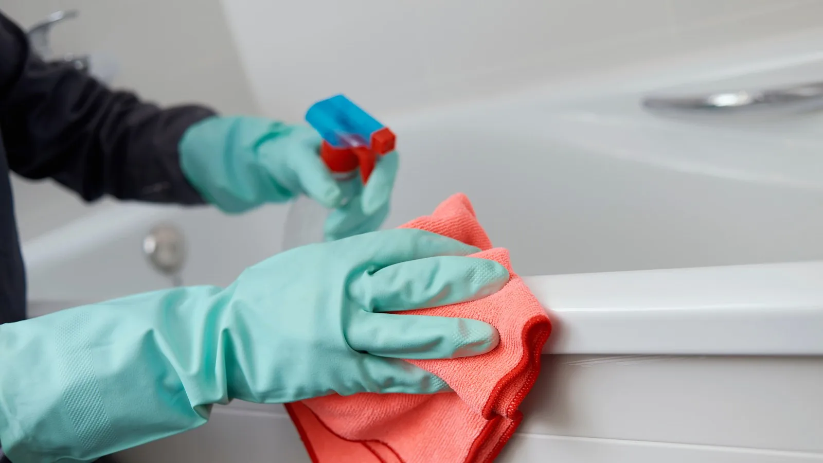 Gloved hands cleaning a bath surround with a trigger spray and cloth.